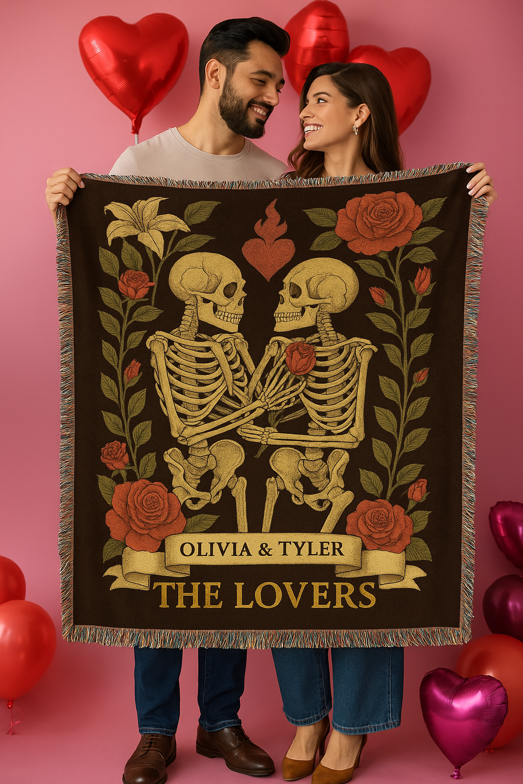 Two people holding a blanket with skeletons and roses design against a pink background with heart balloons.